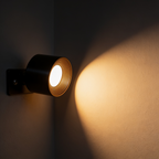 Aurevia cordless LED wall sconce, black cylindrical head on a wall plate, casting a warm elliptical pool of light across a plaster wall at night
