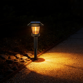 Black solar pathway light with a visible warm amber filament bulb, staked beside a gravel garden path and pooling light on the stones