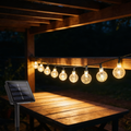 Solar crystal globe string lights strung along a garden pergola rail above a wooden table, warm white bulbs glowing at night