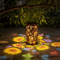 Hanging rust-brown metal solar lantern projecting coloured sunflower and butterfly patterns across a stone garden patio at night