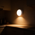 Round white motion sensor puck night light mounted on a kitchen wall, washing warm light down onto the worktop below