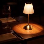 Cordless pleated table lamp glowing warm on a dark walnut restaurant table beside a wine glass and cutlery, no cable