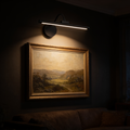 The Pivot cordless picture light mounted above a framed landscape painting in a dark room, washing warm light down over the canvas