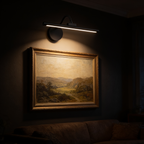 The Pivot cordless picture light mounted above a framed landscape painting in a dark room, washing warm light down over the canvas